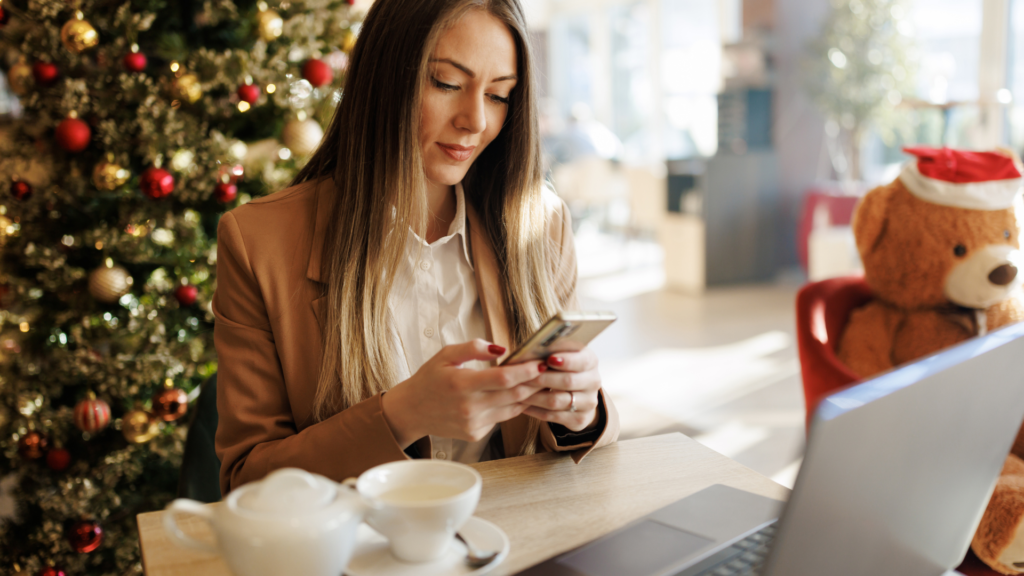 woman on her phone and laptop while the background has christmas tree and holiday decor