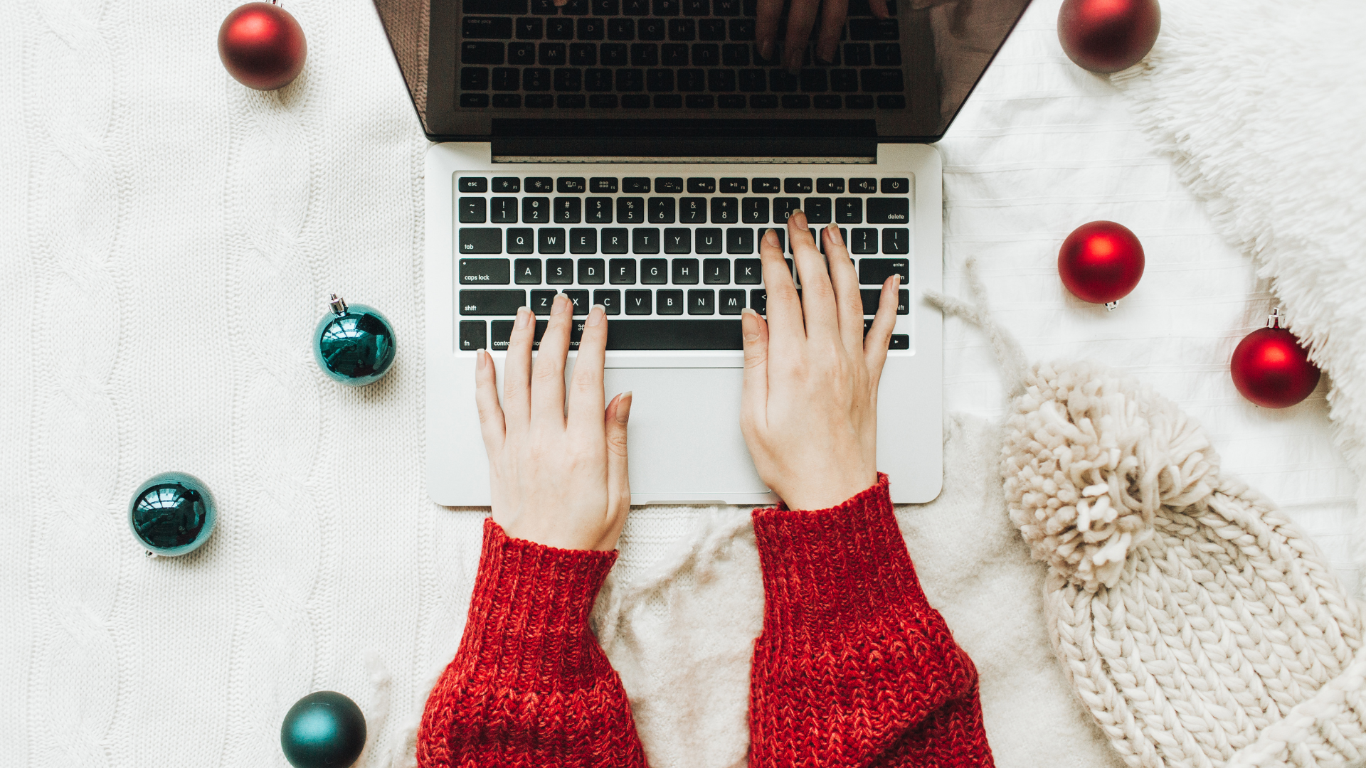 woman wearing red sweater and working on laptop while there are christmas ornaments around it and winter hat